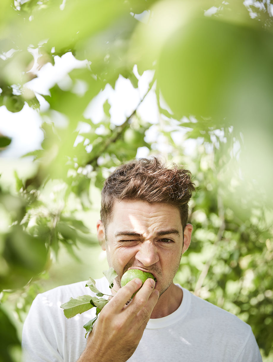 Ralph Schelling beisst in einen grünen Apfel, während er unter einem Apfelbaum steht und ein weisses T-Shirt trägt.